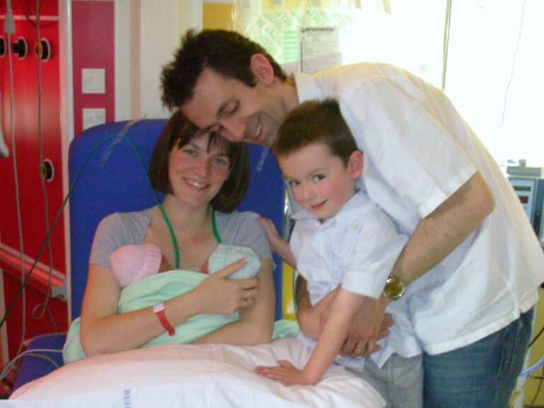 A woman sits on a hospital bed holding a newborn wrapped in a blanket. A man and young boy stand close, smiling at the baby and the camera—capturing the perfect blog post moment in a cosy hospital room.