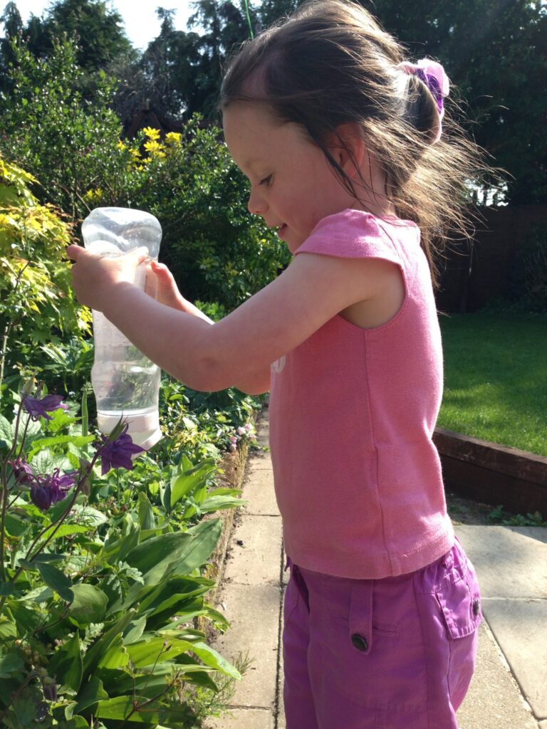 A young girl in a pink top and purple shorts waters plants in a garden on a sunny day, smiling as she uses a ketchup bottle. Lush greenery and flowers surround her, adding to the fun atmosphere.