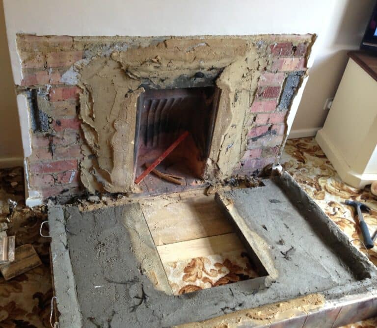 A brick fireplace under renovation, featuring unfinished plaster and exposed bricks, hints at a modern fireplace transformation. Tools rest in the hearth atop a concrete base with a rectangular cut-out, while a patterned carpet surrounds this evolving space.
