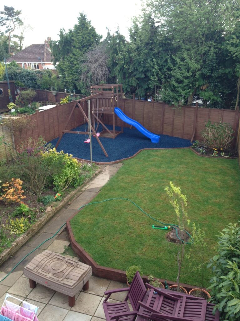 A back garden renovation featuring a lush lawn, vibrant flower beds, patio furniture, a hosepipe, and a wooden playset with swings and a blue slide on blue rubber mulch, all surrounded by a wooden fence and mature trees.