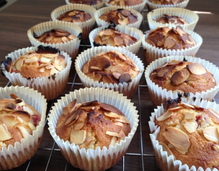 Freshly baked almond muffins, reminiscent of delicate almond cakes, are topped with flaked almonds and cooling on a metal rack. The muffins, in white paper cases, are arranged closely together on a wooden surface.