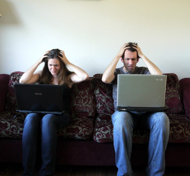 You and I sit on a sofa with laptops, looking frustrated and holding our heads with both hands, suggesting we're experiencing stress or difficulty while using our computers.