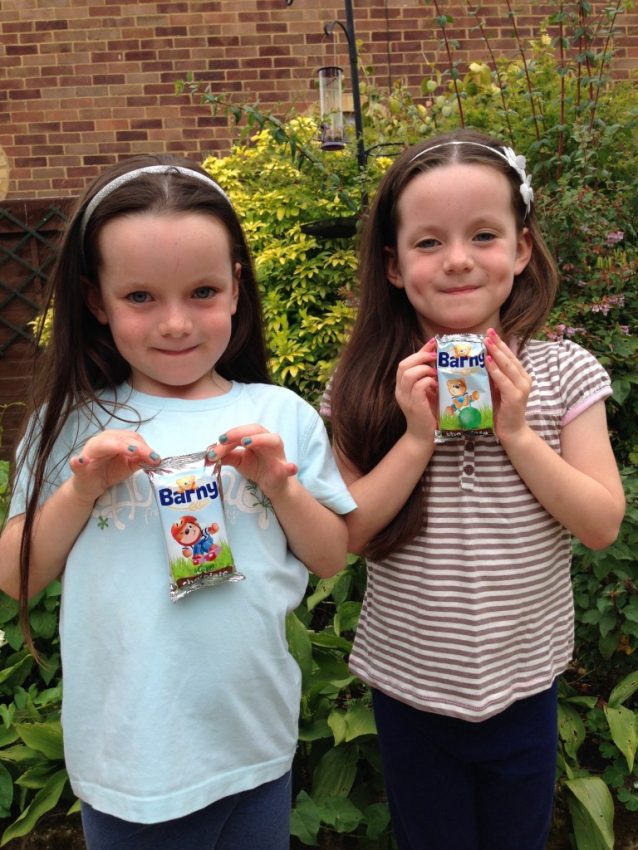 Two young girls with long brown hair stand outside in a garden, each holding a Barny Bear snack cake. Smiling and close together, they look ready for carnival adventures amid leafy green plants and a brick wall in the background.