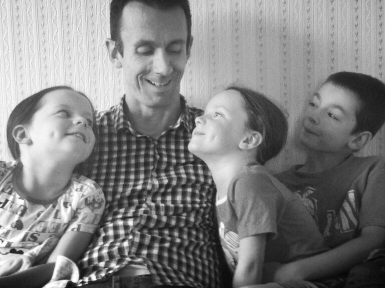 A man sits smiling with three children, two girls and one boy, all looking at him affectionately. This black and white dad photo captures a joyful moment—perfect as a heartfelt gift to celebrate family togetherness.