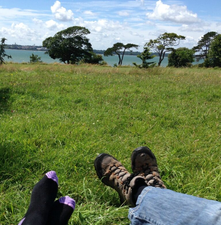 You and I relaxing on the grass in July, gazing at a scenic lake and distant trees; one in walking boots and jeans, the other in black socks with purple toes. A blue sky with scattered clouds stretches overhead.