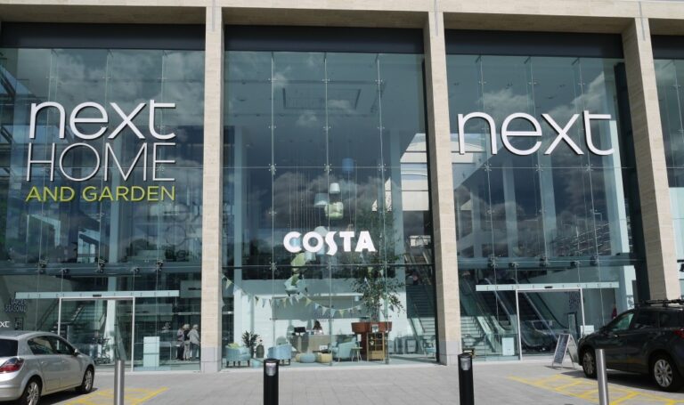 Glass-fronted building featuring large NEXT Hedge End HOME AND GARDEN signs, a prominent COSTA coffee shop inside, and cars parked outside. This Southampton superstore offers a modern shopping experience.
