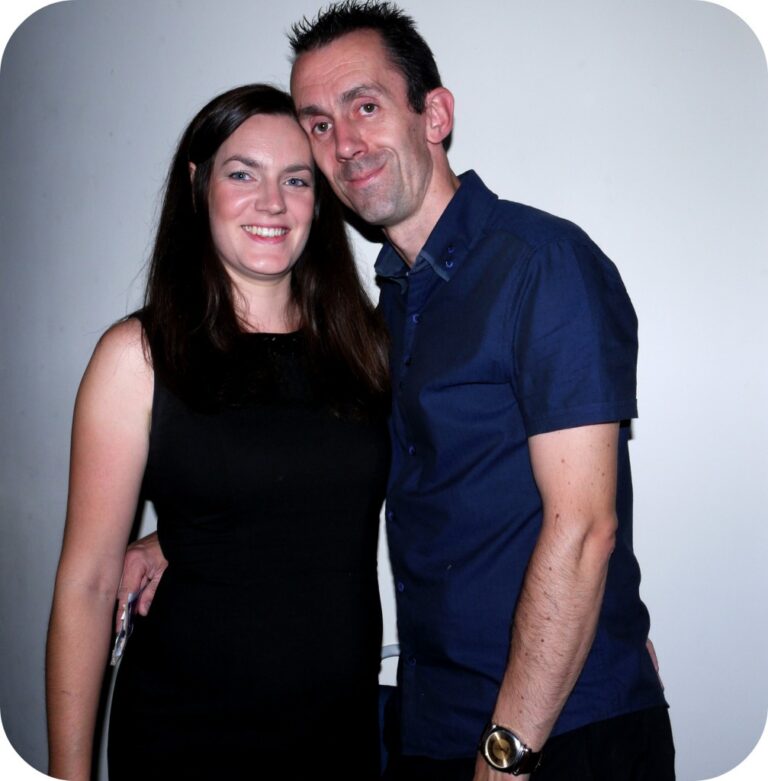 You and I, smiling together in style—a woman in a black dress and a man in a navy blue shirt—stand close against a plain white background, capturing an August moment.