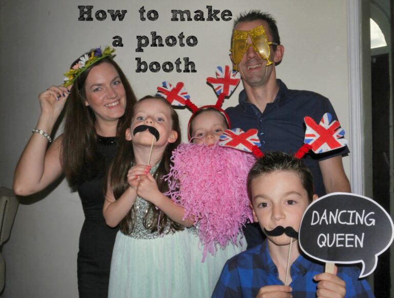 A smiling family poses for a DIY birthday photo booth, wearing fun props like fake moustaches, a pink beard, novelty glasses, flower crown, and British flag headbands. A sign reads DANCING QUEEN and text above says make your own photo booth.