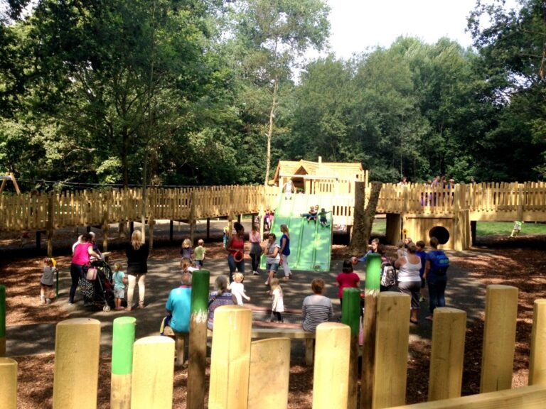 Children and adults gather at the adventure playground in Royal Victoria Country Park, enjoying wooden structures, slides, and climbing equipment surrounded by trees and greenery on a sunny day.