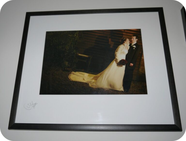 Framed photograph of a bride and groom standing together outdoors at night, both smiling in formal wedding attire. Perfect for creating a family wedding photo wall, the image is mounted with a white mount and displayed in a black frame.