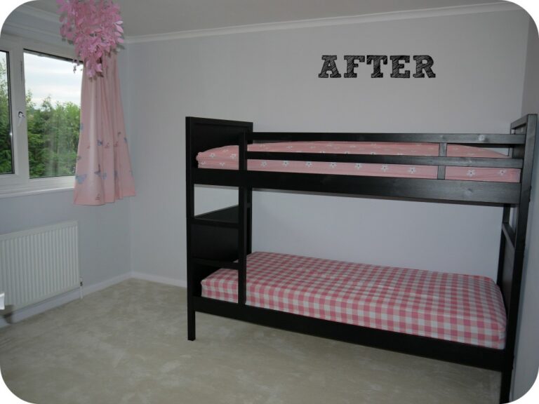 A clean, minimal twin girls' bedroom with a black bunk bed; both mattresses have pink chequered sheets. The wall is light grey, featuring “AFTER” in black letters. A window and a pink decorative light fitting complete this bedroom renovation.