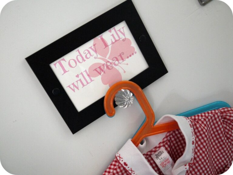 A framed sign on the wall reads, Today Lily will wear... in pink text with a butterfly. Below, a red and white checked shirt—perfect for kids' outfit ideas—hangs on an orange hanger with a clear decorative hook.