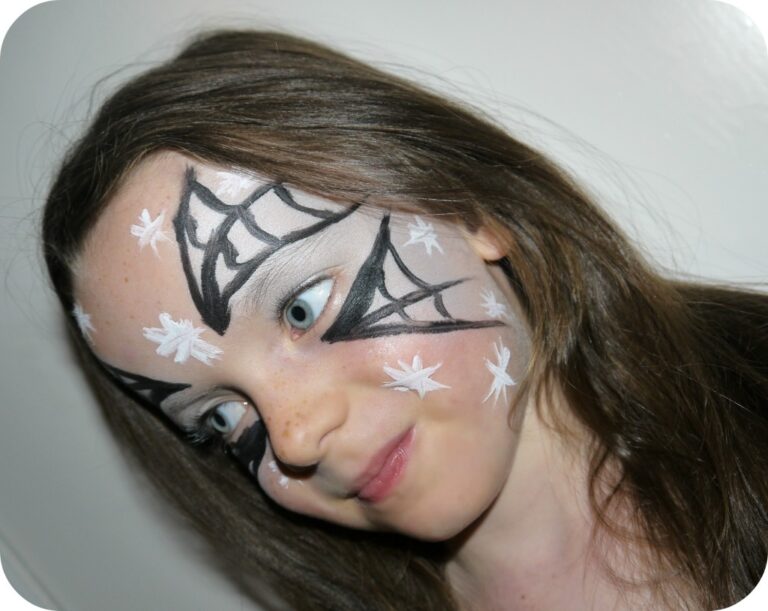 Young girl with long brown hair and light skin has her face painted with black and white spider’s web designs around her eyes and white stars on her cheeks, smiling gently—a perfect look for Hallowe’en face painting.