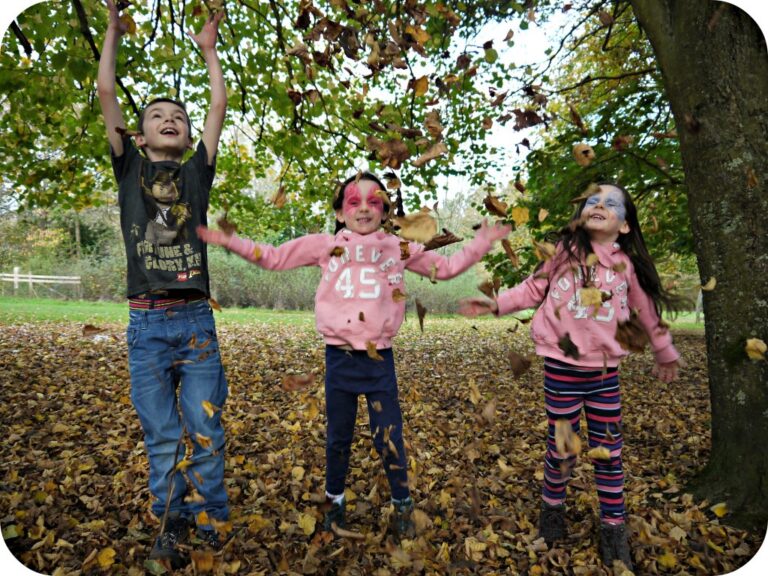 Three children with face paint have fun as they joyfully throw autumn leaves into the air whilst standing under a tree in a park, surrounded by fallen leaves and greenery.