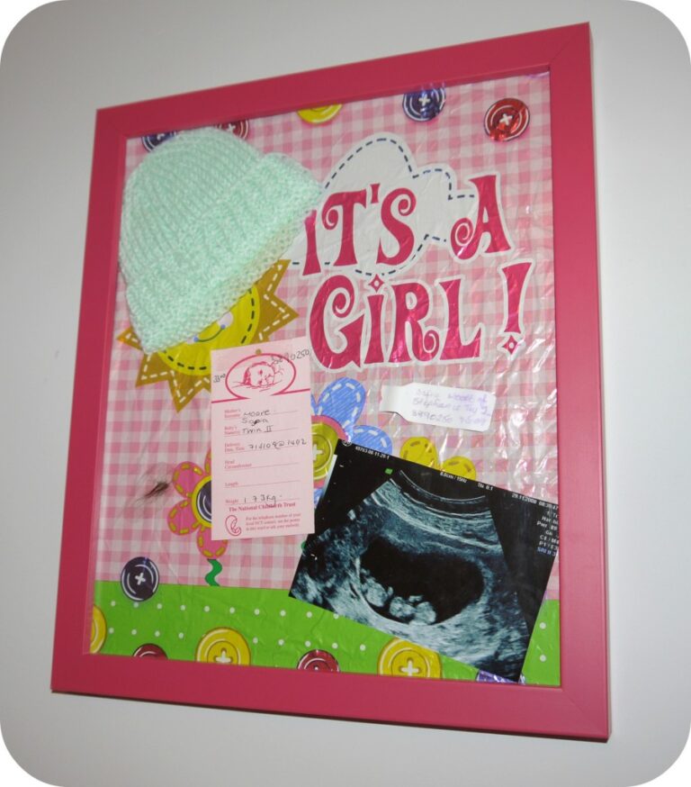 A pink framed collage, perfect as a baby keepsake, features a sign reading "It's a Girl!", a small mint green knitted hat, hospital card, and black and white ultrasound photo on a pink chequered background.