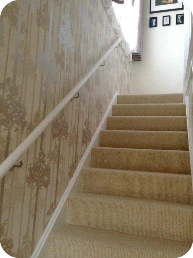 Carpeted staircase with a white handrail on the left, beige wallpaper featuring a tree pattern, and framed pictures at the top of the stairs. Light from a window above brightens the finished hall.