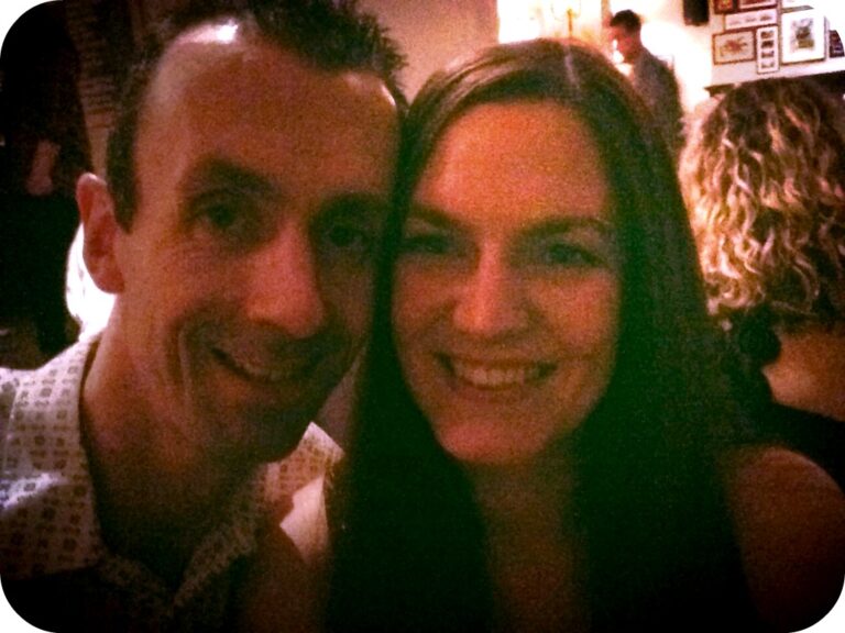 You and I are smiling and posing closely together for a photo in a dimly lit indoor setting, with other people and framed pictures visible in the background—capturing a memorable November moment.