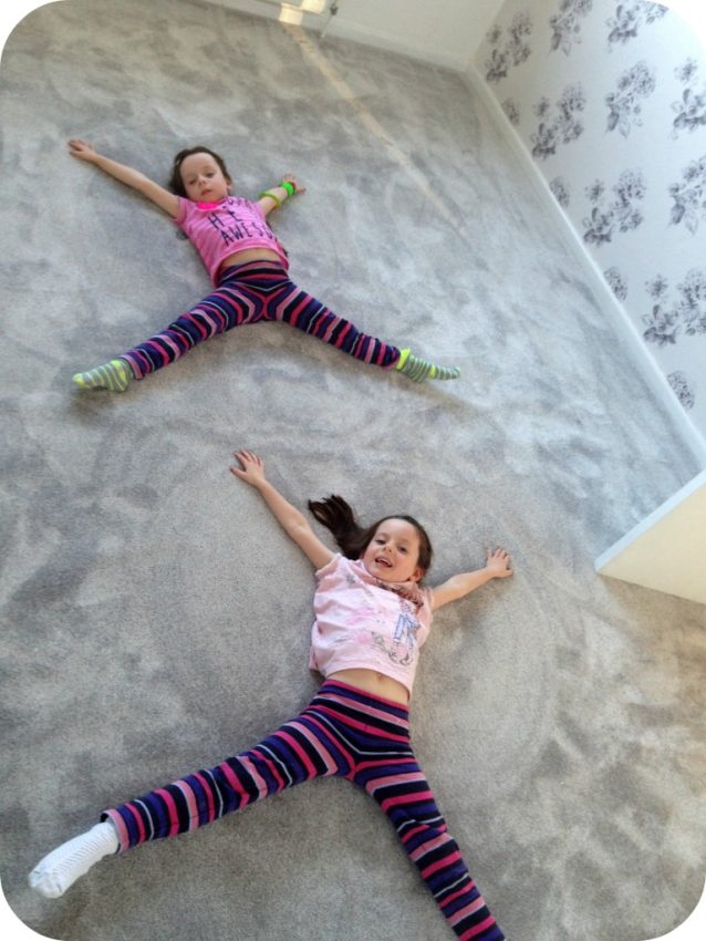 Two young girls lie on a light grey carpet in a freshly completed bedroom renovation, arms and legs stretched out in a star shape, smiling and wearing matching striped trousers and pink tops in a brightly lit room.