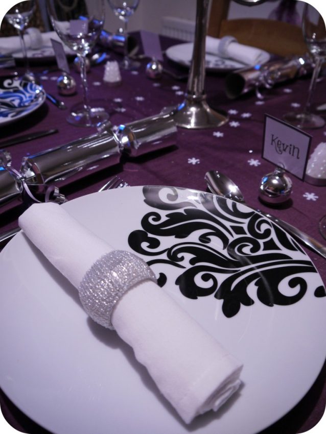 A close-up of an elegant Christmas table setting with a white plate featuring a black decorative pattern, a white serviette in a silver serviette ring, and a place card labelled Kevin on a purple tablecloth for festive table inspiration.