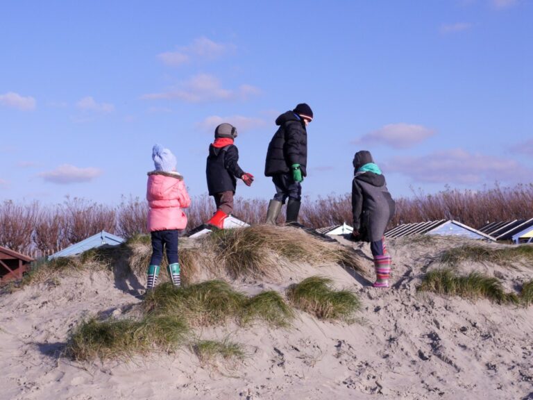 Four children in winter clothing and boots stand on a sandy dune with grass at West Wittering beach, each facing a different direction. Beach huts and tall grass appear in the background beneath a blue sky.