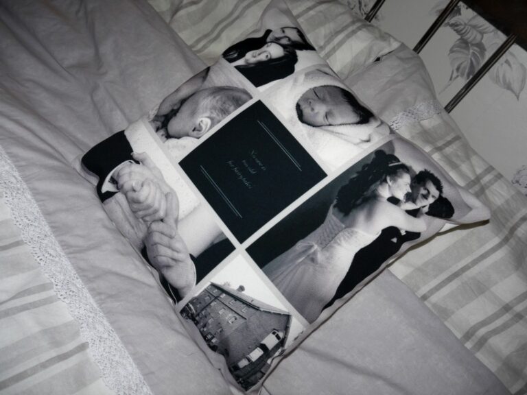 A PhotoBox story photo cushion featuring black-and-white snapshots—a newborn, wedding day couple, two hands, and a house—rests on a neatly made bed with striped and lace-trimmed bedding. A unique Valentines gift for cherished memories.