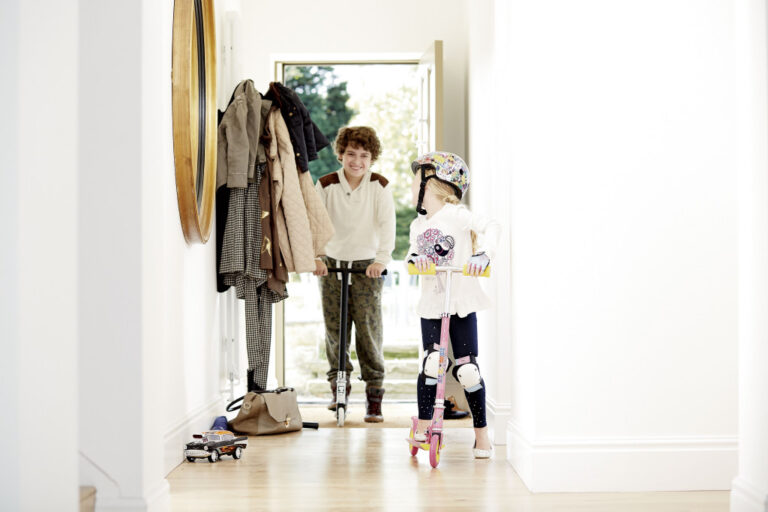 Two children are in a bright hallway of their home; one girl wearing a helmet and pads rides a scooter whilst a boy stands behind her with a scooter by the open front door. Plaster walls and coats hang on hooks to the side, adding charm to Rooms Made For You.