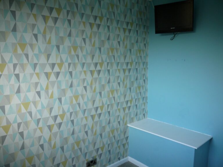 A boy's bedroom corner featuring a geometric-patterned feature wall in shades of blue, grey, and yellow, a pale blue wall, a white bench, and a small flat-screen TV mounted on the wall.
