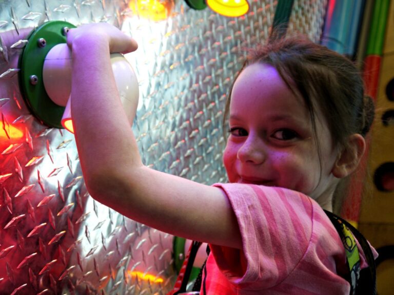 A young girl in a pink shirt smiles at the camera while pressing a large, round button on a colourful, illuminated metal wall at Rock Up Whiteley, capturing the fun atmosphere perfect for any Rock Up Whiteley review.