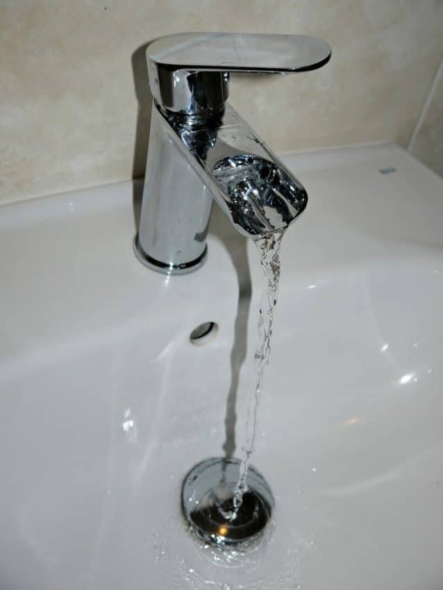 A modern chrome Arte Basin mixer tap with water flowing into a white bathroom basin. The basin waste is visible below the stream of water. Perfect for a Soakology bathroom setup—discover more in our review. Tiled background adds a sleek finish.