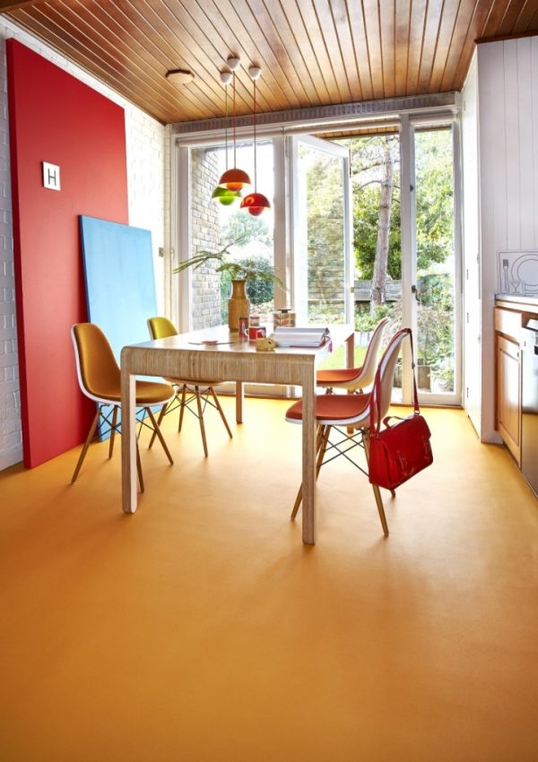 A modern dining area with a yellow floor, unique carpet, wooden table, four colourful chairs, and hanging multicoloured lamps. Large glass doors open to a green garden, while red and blue accents add vibrancy. A red bag hangs on a chair.