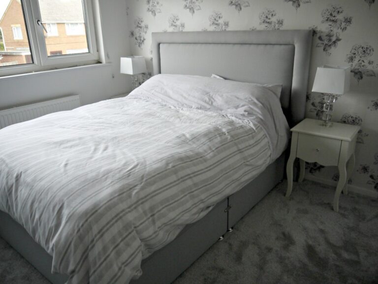 A neatly made bed with a light grey headboard and striped bedding, possibly an Ottoman bed, sits between two white bedside tables topped with lamps, in a carpeted room with floral wallpaper and a window on the left—perfect for a Dreams review.