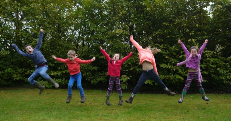 Five children wearing colourful clothes jump with arms raised on a grassy lawn during an Alresford walk, with green bushes in the background. They appear joyful and energetic, captured mid-air in an outdoor setting.