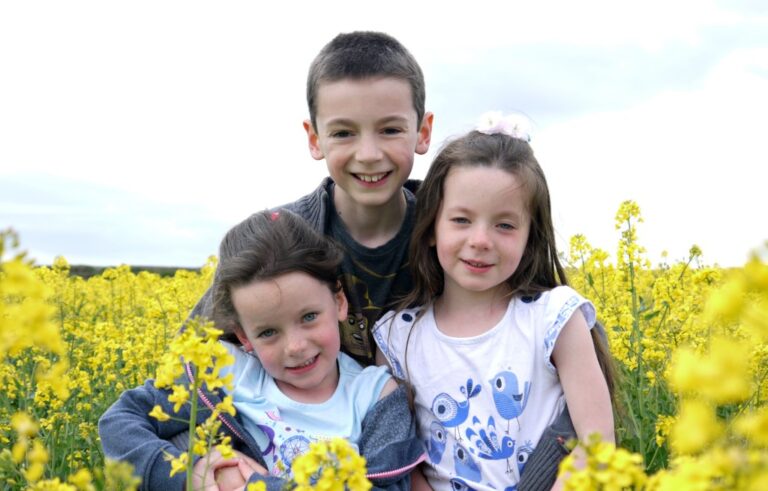 Three smiling children, two girls and one boy, sit closely together in yellow fields under a bright sky. The children look happy and are dressed in casual clothes, surrounded by beautiful yellow flowers.