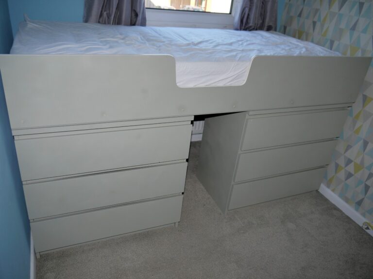 A white loft single bed with a mattress on top, supported by two sets of white drawers in an IKEA MALM hack, sits against a blue and patterned wall in a carpeted bedroom.
