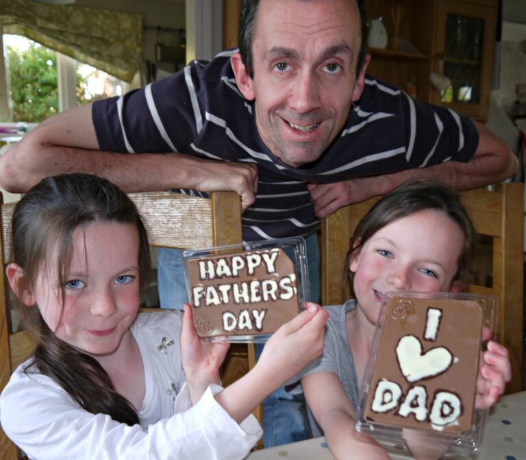 A smiling man stands behind two young girls at a table, each holding up Make-a-Bar chocolate bars with messages: HAPPY FATHER'S DAY and I ♥ DAD. They are indoors, celebrating together.
