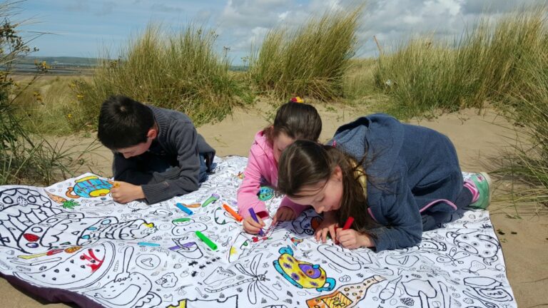 Three children lie on an Eggnogg colour-in picnic blanket spread over sand dunes, colouring a large illustrated sheet with felt-tip pens. Tall grasses surround them, and the scene is bright and sunny under a partly cloudy sky.