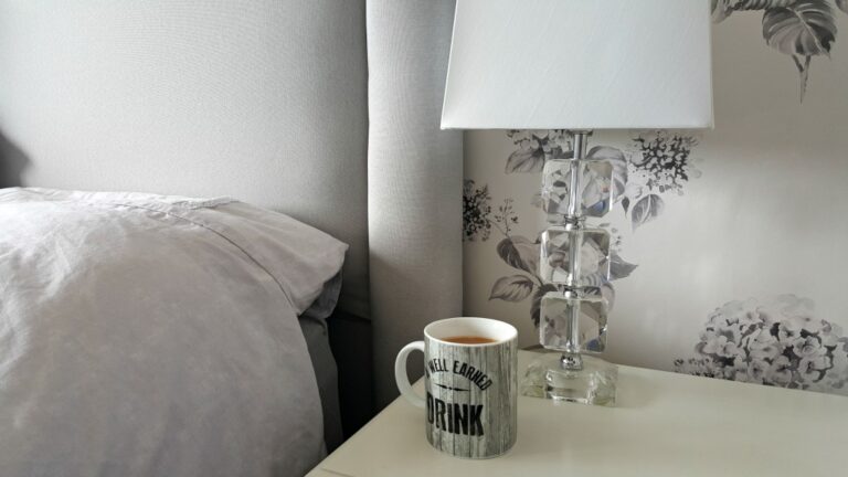 A grey mug with the words “Well Earned Drink” sits on a white bedside table next to a clear glass lamp, with a grey pillow and floral wallpaper in the background—perfect touches to help improve my sleep, inspired by Warren Evans sleep results.