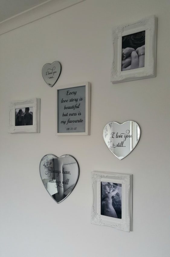 A white wall decorated with six framed black-and-white photos and three heart-shaped mirrors from Best4Frames. One rectangular frame in the centre displays a romantic quote about love. Enter their competition for a chance to win £50! The arrangement forms a loose cluster.