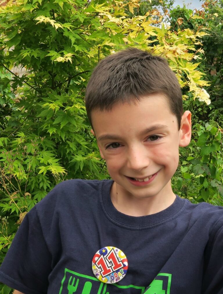 A young boy with short brown hair smiles at the camera whilst standing outdoors in front of green foliage. He is wearing a dark T-shirt and a colourful badge that says 11 Today, ready for his 11th birthday celebration.