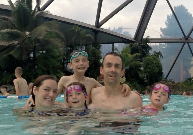 A family of five, wearing swimming goggles, smiles whilst standing together in a tropical indoor swimming pool at Center Parcs Sherwood Forest. Lush plants and trees surround the pool, creating a warm and inviting atmosphere perfect for a memorable video blog.