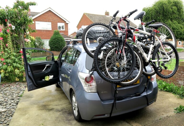 A silver car parked in a driveway has its rear loaded with three bicycles on a bike rack, ready for a Centre Parcs Sherwood Forest adventure. The car’s boot is open, with houses and greenery visible in the background.