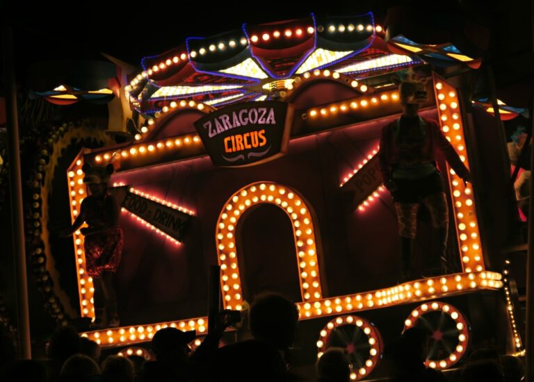 A brightly lit circus float reading Zaragoza Circus shines at night during Bridgwater Carnival 2015, with two performers in costume standing on either side, surrounded by colourful lights and silhouettes of onlookers.