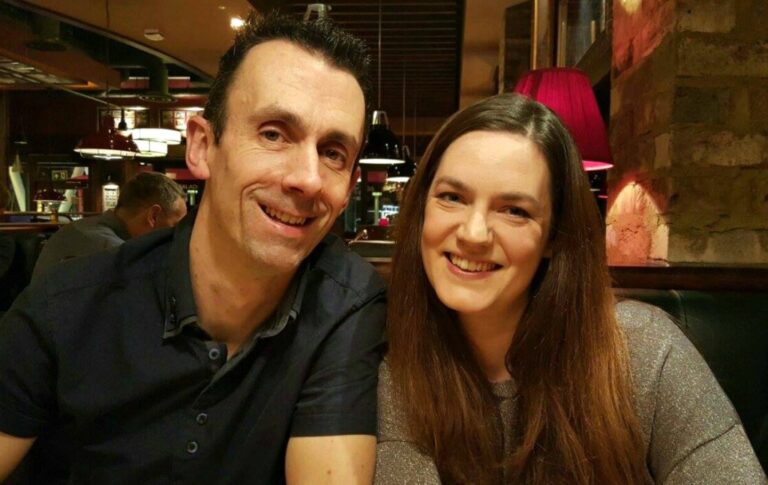 A man and woman sit closely together in a warmly lit restaurant at Coast 2 Coast Whiteley, smiling at the camera. Red lamps and wooden decor in the background create a cosy atmosphere.