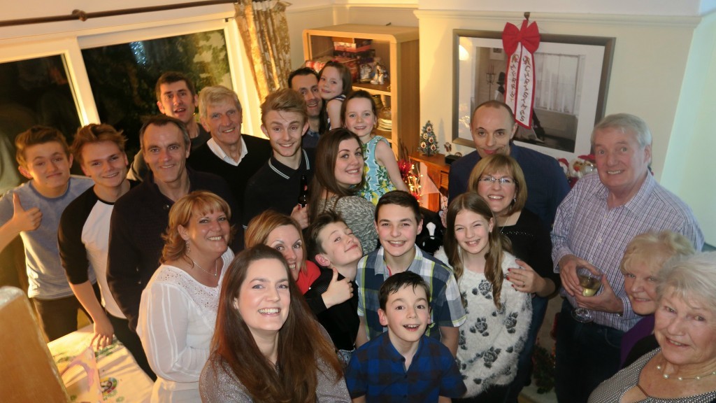 A large group of smiling people of various ages pose closely together indoors, likely celebrating a festive occasion. With festive decorations and a small Christmas tree in the background, this moment could be part of a completed Project 365 photo-a-day series.