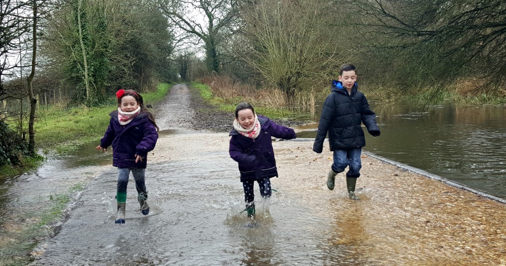 Three children wearing coats and wellies joyfully run through a shallow, flooded path along the Titchfield Canal in a rural area, surrounded by bare trees and greenery on a cloudy day—a classic wet walk adventure.