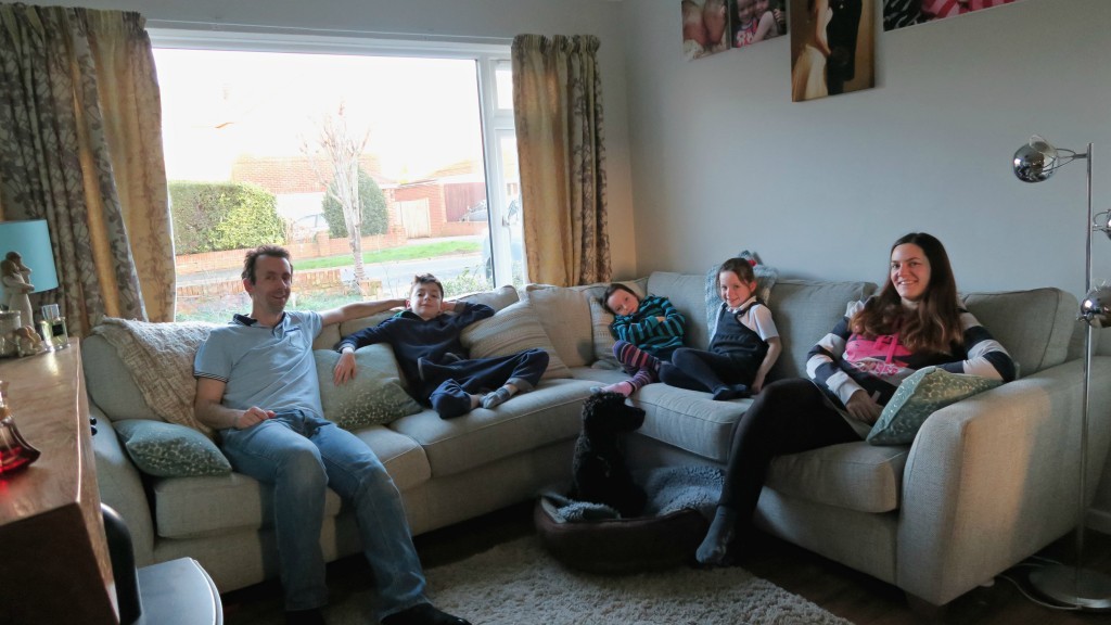 A family of five sits on a beige corner sofa in the lounge, smiling and relaxing together. Two dogs join in the love—one sits with the family whilst the other naps contentedly in a dog bed on the floor.
