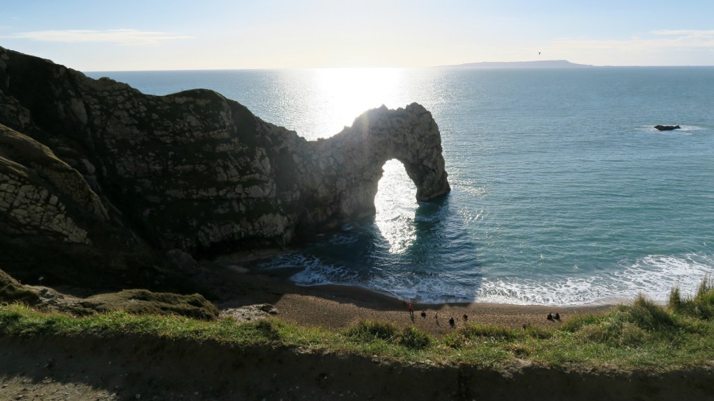 Durdle Door, the iconic rocky arch on the Jurassic Coast in Dorset, stands tall as sunlight glistens on the sea, waves crash on the sandy beach, and a few people stroll along the shore.