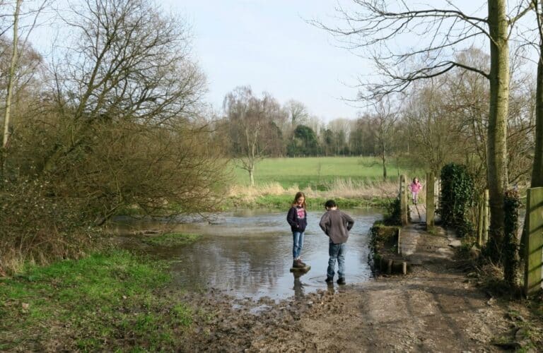 Two children in kids' walking boots stand by a shallow, muddy stream on a rural path surrounded by bare trees and green fields. Another person walks in the background. The scene is overcast and peaceful.