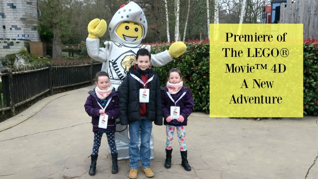Three smiling children stand in front of a person in a LEGO knight costume outdoors. A yellow sign reads: “Premiere of The LEGO Movie 4D: A New Adventure.” Trees and buildings complete the fun LEGO Movie scene.