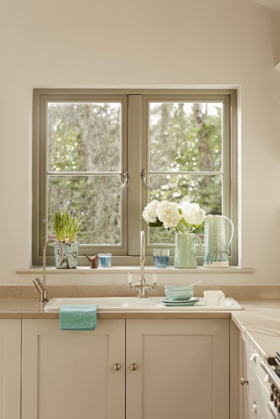 A tidy kitchen with wooden windows above the sink displays potted flowers, a green jug, and a blue glass jug. Light cupboards, a folded turquoise towel, and neatly arranged crockery create a calm, inviting makeover.